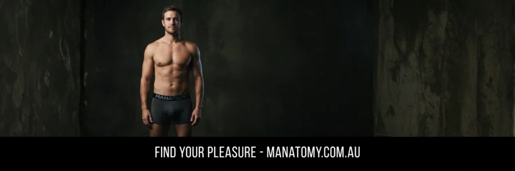 A man in grey Manatomy trunks standing against a dark stone wall, above a Find Your Pleasure banner.